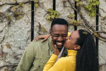 A laughing man in an olive jacket as a woman in a yellow sweater kisses his cheek beside an ivy-covered wall