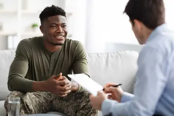 A smiling man in an olive shirt and camouflage trousers talking with a clinician taking notes