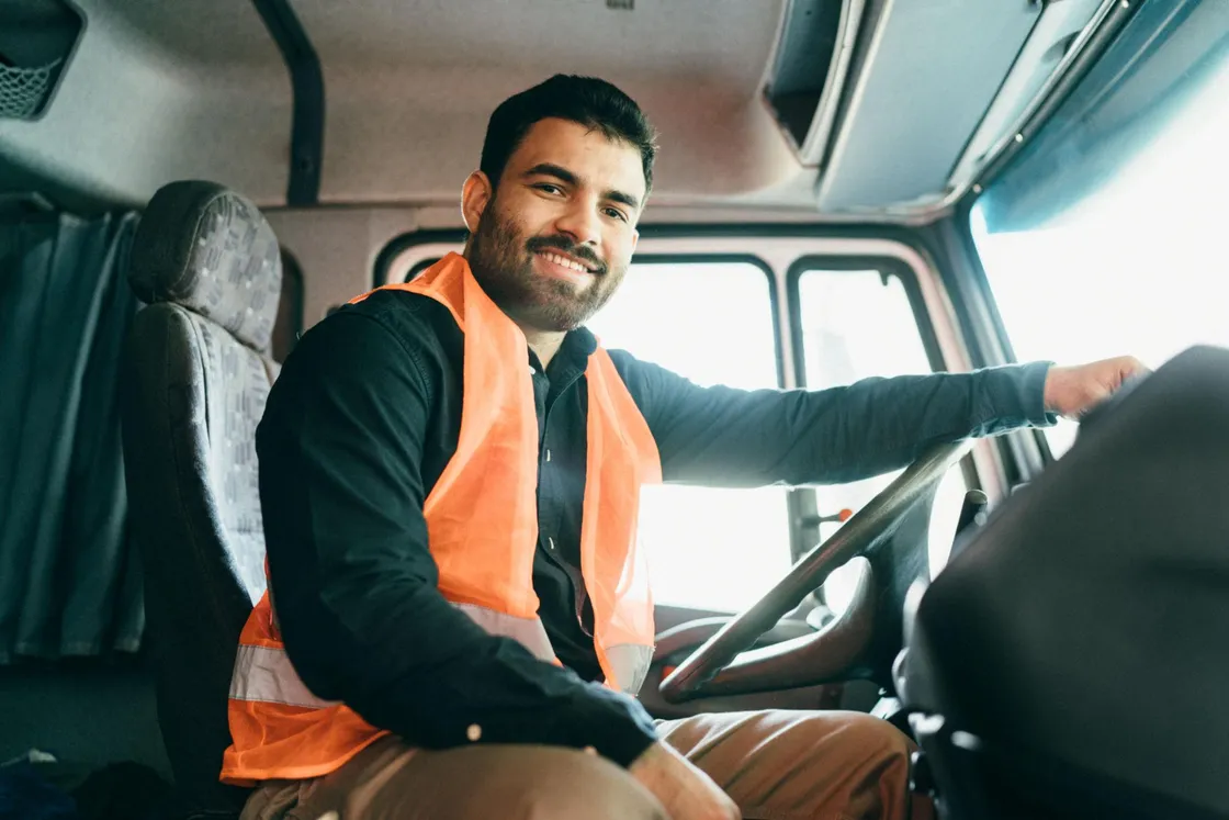 A smiling commercial driver in a hi-vis vest at the wheel of a truck cab