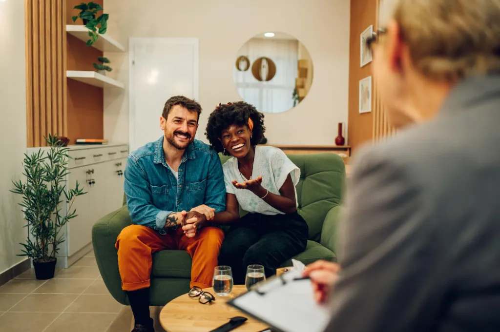 A couple sitting together on a sofa in a consulting room, smiling as they talk with a therapist