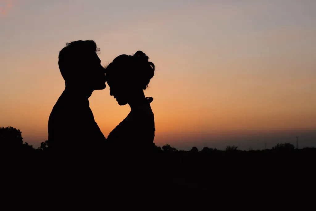 A couple silhouetted against an orange sunset sky, standing close with their foreheads almost touching