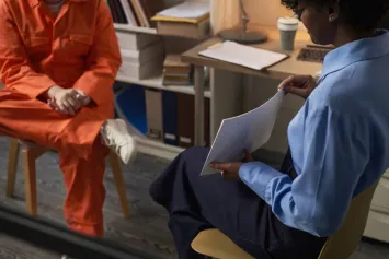 An evaluator in a blue shirt reading a document opposite a person seated in an orange jumpsuit