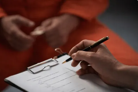 An evaluator writing notes on a clipboard opposite a man in an orange jumpsuit