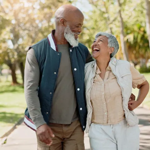 An older couple walking together outdoors in a park, laughing as they look at one another