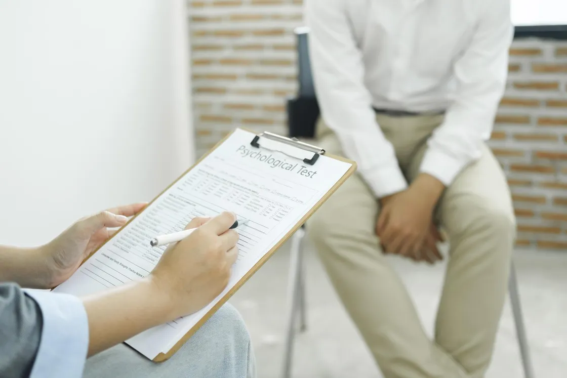 A clinician completing a psychological test form on a clipboard while a seated man waits