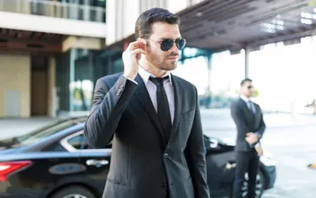A security professional in a dark suit, tie and sunglasses touching his earpiece outside a building, a black car behind him