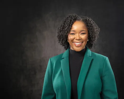 Dr. Simone Wright in a green blazer and black top, smiling, against a dark grey backdrop