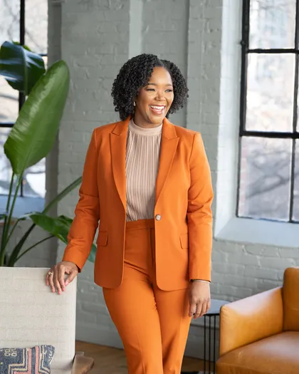 Dr. Simone Gillon-Wright in an orange suit, laughing and looking away, one hand resting on a chair