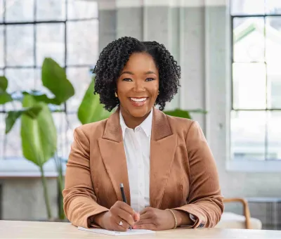 Dr. Gillon-Wright in a tan blazer smiling at the camera while writing in a notebook at a desk