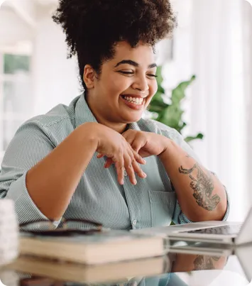A smiling woman resting her chin on her hands at a desk beside an open laptop and a stack of books