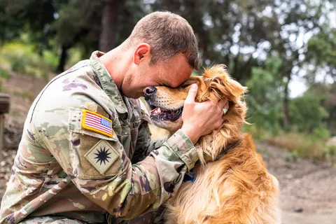 A soldier in camouflage kneeling outdoors, resting his forehead against a golden dog he is holding