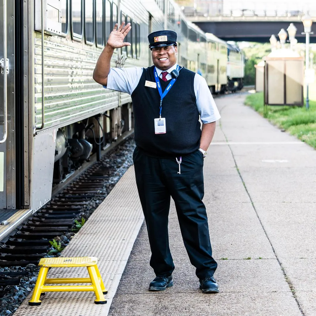 A smiling train conductor waving beside a passenger train on the platform