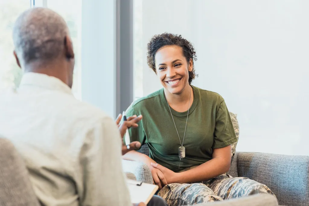 A young woman veteran in an olive T-shirt and dog tags smiling in conversation with a therapist
