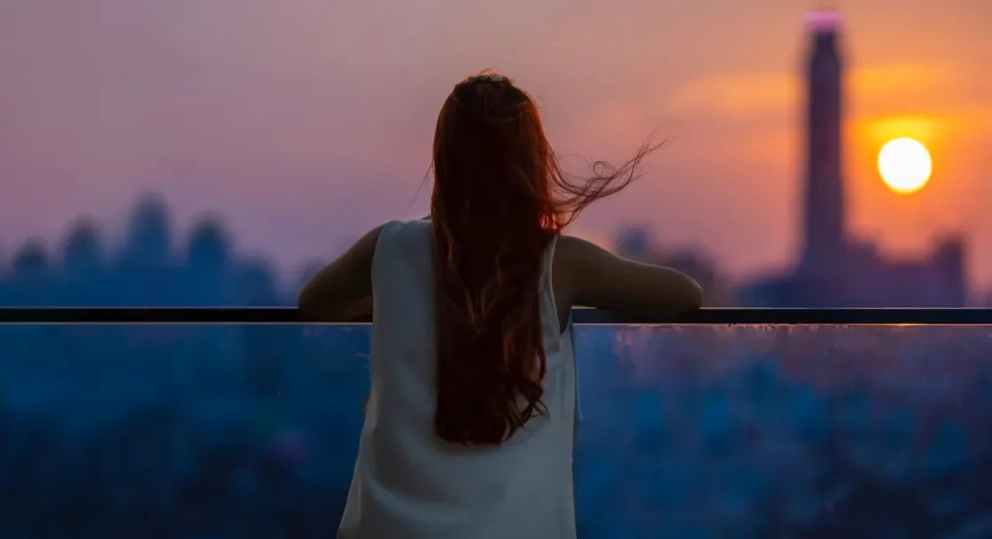 A woman leaning on a balcony rail, looking out over a city skyline at sunset.