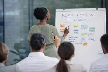 A woman in a green dress writing on a whiteboard headed Mental Health in the Workplace while a group watches