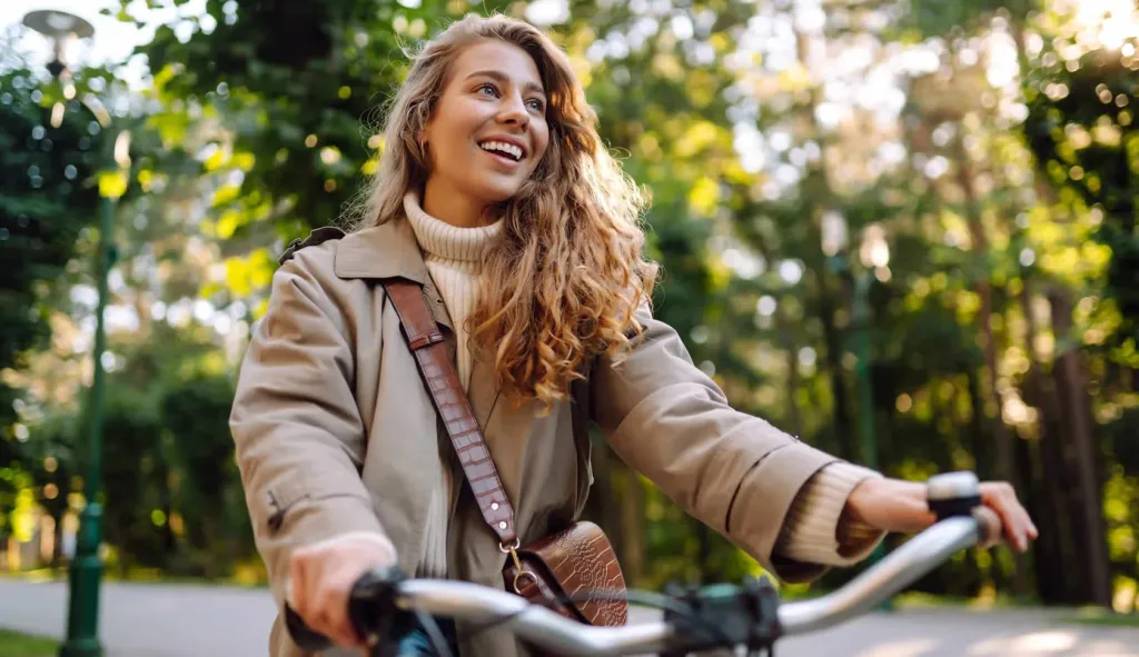 A woman smiling as she cycles along a tree-lined path on an autumn morning.